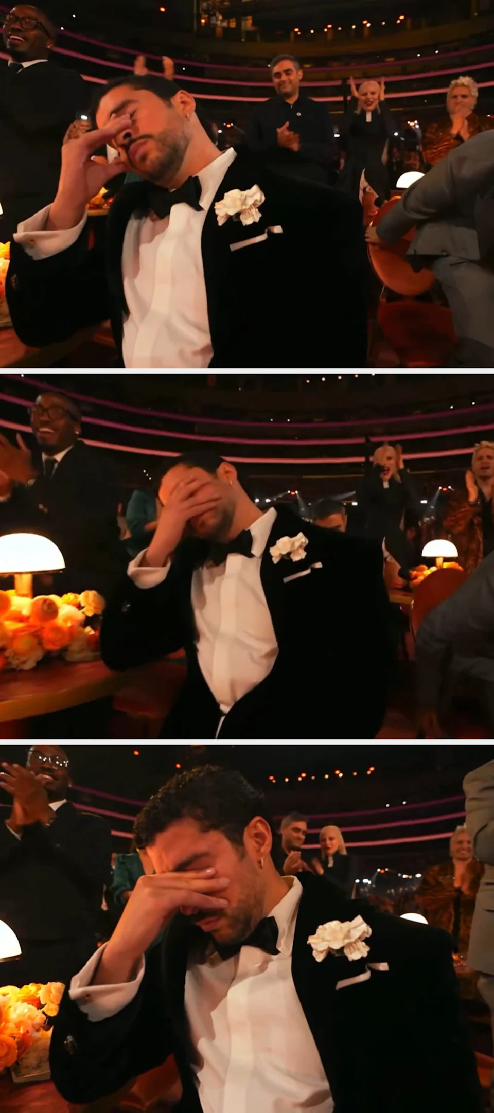 Man in a tuxedo with a white boutonnière, sitting with eyes closed, as people behind him stand and cheer in a formal event setting