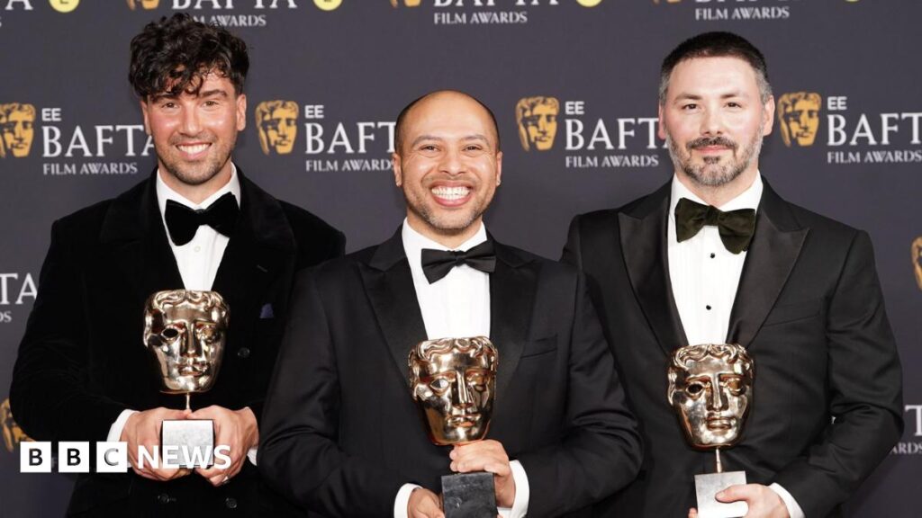 Three men stand on the 2026 BAFTA red carpet in black tuxedos. Each of the three hold a BAFTA award in the shape of a gold theatrical mask.