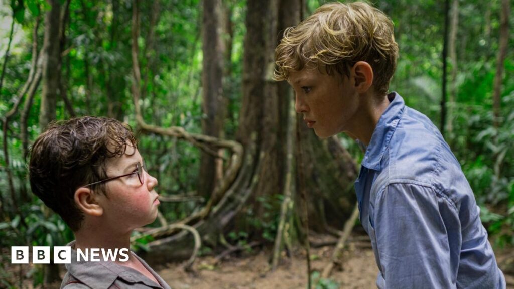 A young boy with brown hair and glasses looks up at a blonde haired boy in a blue shirt. They are in a wooded area of a tropical island.