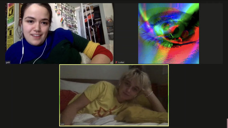 Kami Hinds as Gabi in a blue shirt with yellow and red sleeves laying on her bed in her room talking to Mikhala Hoffman as Levi, also laying on their bed, in a yellow and red t-shirt, with a third person in the chat with the camera off and their multicolored eye icon on display in Concrete Paradise