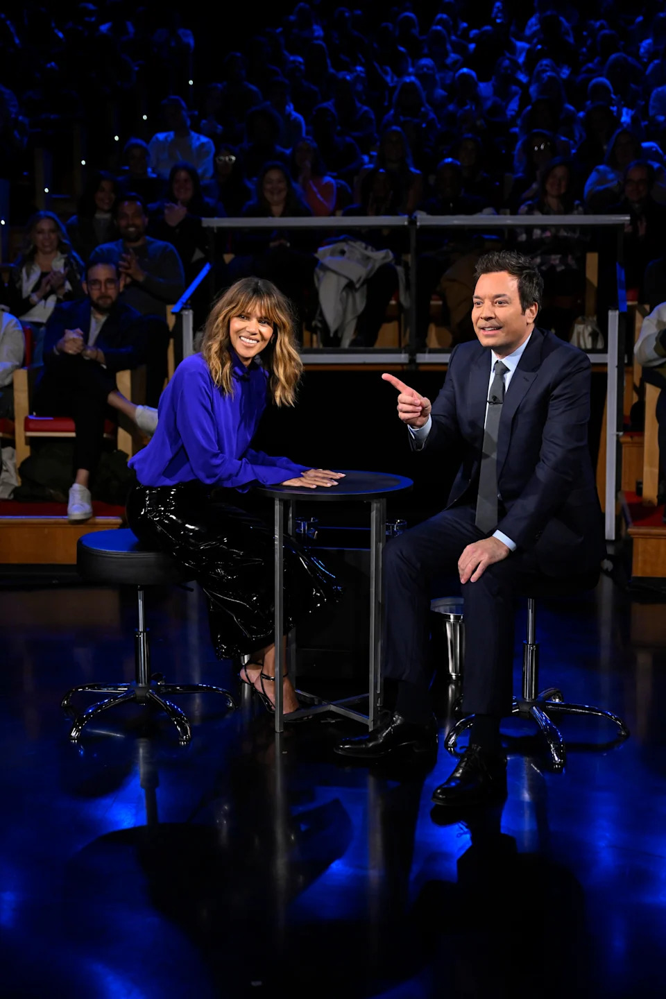 Halle Berry in a chic outfit and Jimmy Fallon in a suit sit at a table on a talk show set, smiling and engaging with the audience