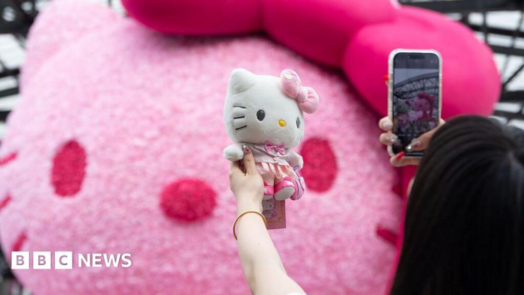 Hello Kitty designer steps down after 46 years A woman holds up a Hello Kitty soft toy to take a photo with her phone. Behind the toy there is a large pink Hello Kitty head