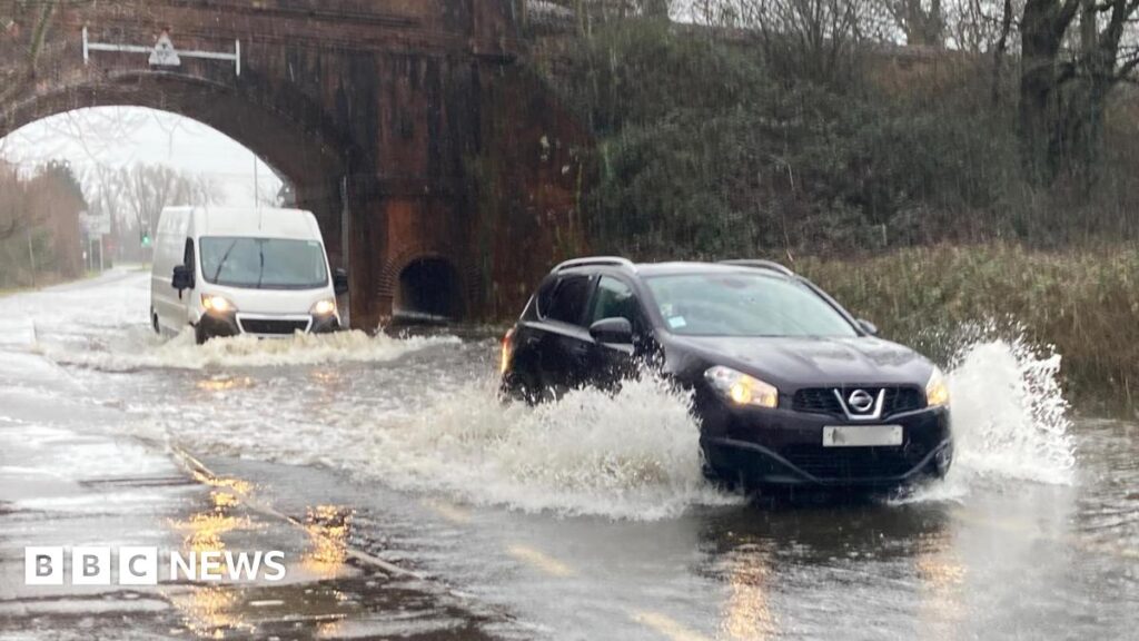 A car and a van drive through a deep puddle under a bridge, spraying water.