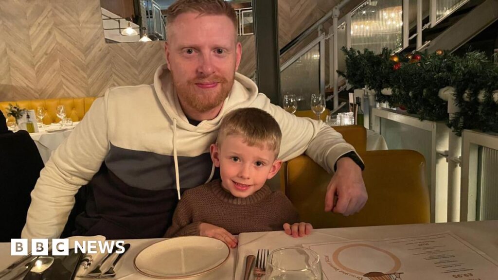 Dad of deaf Coronation Street star to tackle Everest Michael Woods, 36, who has ginger hair and a short ginger beard, sits at a table in a restaurant with his arm around his son, six, who has short blonde hair.