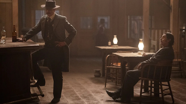 Seth Bullock (Timothy Olyphant) stands at a bar while Al Swearengen (Ian McShane) speaks to him while seated at a table in Deadwood: The Movie