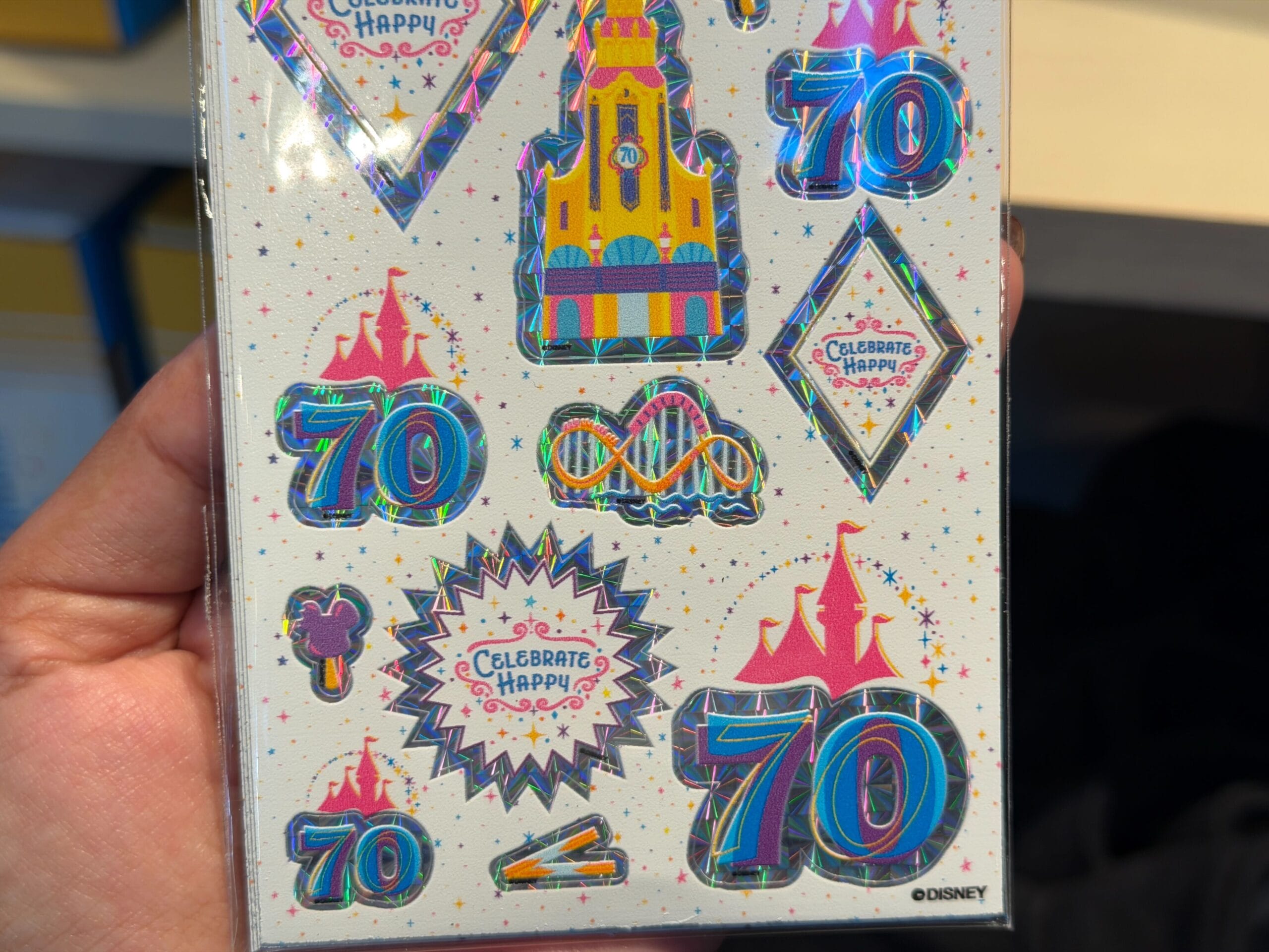 The stickers on the visible sheet feature holographic edges and designs like the 70th anniversary logo, churros, Mickey ice cream bars, 