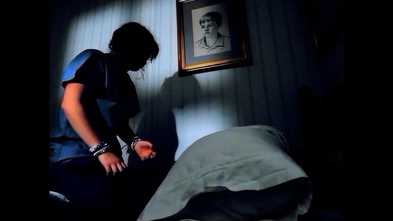 Betsy Sligh as Monroe kneeling on a bed in shadow with a framed drawing of a young boy on the wall and a pillow in view in Dooba Dooba