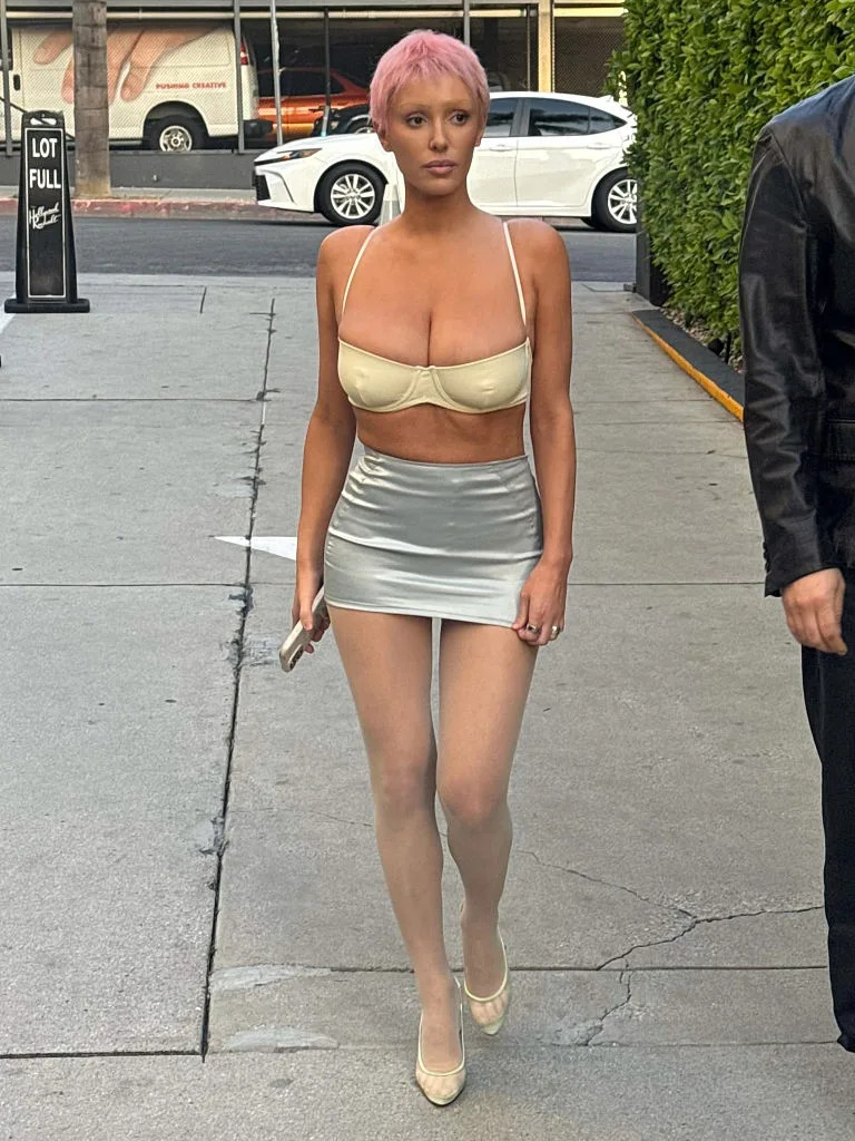 Person on sidewalk wearing a revealing outfit with a crop top and a short skirt, holding a phone