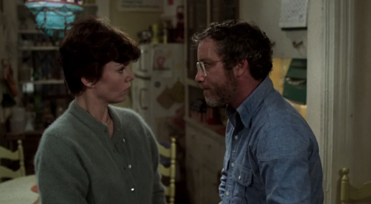 A woman in a green sweater and a man in glasses and a blue shirt stand facing each other in a kitchen, both appearing serious and engaged in conversation. The kitchen is cluttered with various items in the background.