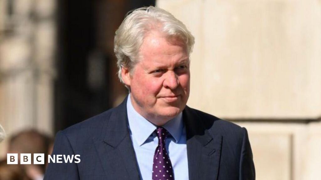 Charles Spencer wears a blue suit, blue shirt and purple tie with white spots in it. He is outside, by a large building. He is looking away from the camera.