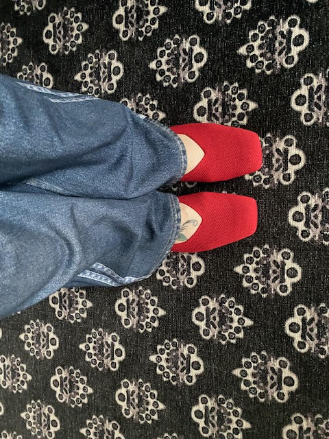 A person wearing blue jeans and red slip-on shoes on a dark patterned rug.