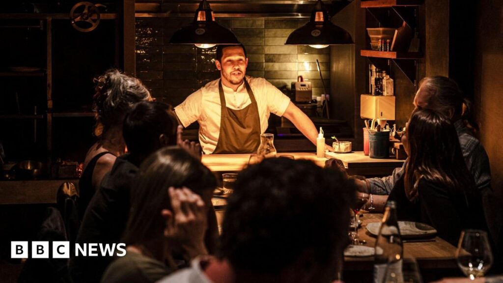 Chef Corrin in kitchen which is open to dining area talking to diners at two tables together. The setting is dark with warm overhead lighting and the diners are all looking at him.
