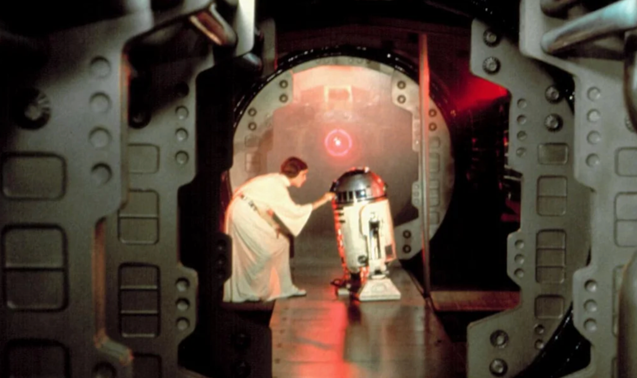 A woman in a white gown bends down to interact with a small blue and white robot inside a metallic spaceship corridor lit by red and yellow lights.