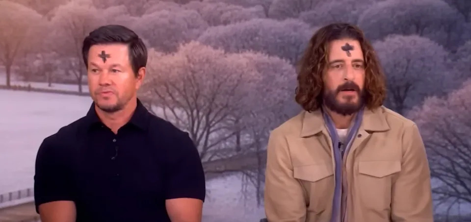 Two men with ash crosses on their foreheads sit for an interview. One wears a black shirt, and the other a beige jacket with a scarf