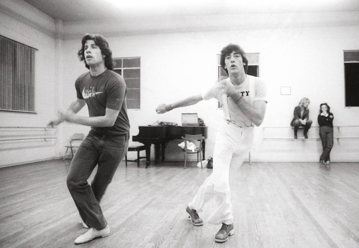 Two men are dancing energetically in a studio with wooden floors; a piano is in the background, and two women sit on benches against the wall, watching them. The scene has a candid, lively atmosphere.