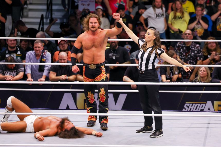 A glowering Adam Page, with beard, mustache, and shaggy hair flowing sideways, stands in the center of a wrestling ring with a referee raising his left arm in the air. Page wears black pants with red-and-gold flame patters on them. To the left of him, sprawled at his feet, Juice Robinson is sprawled on the canvas. Robinson wears white trunks and his hair and beard are shaggy; he is wincing in a show of agony. 