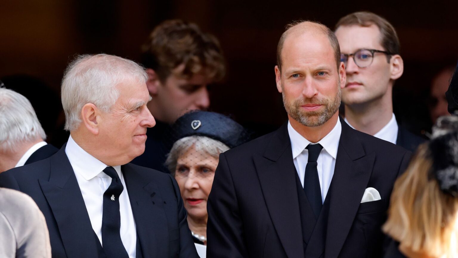Prince William turns to Prince William and smiles at him, while both wear black suits with black ties and white shirts