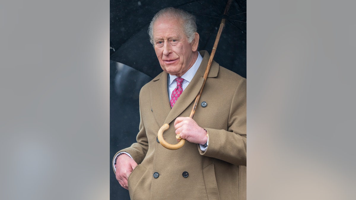 King Charles wearing a beige coat holding an umbrella on a rainy day in the U.K.