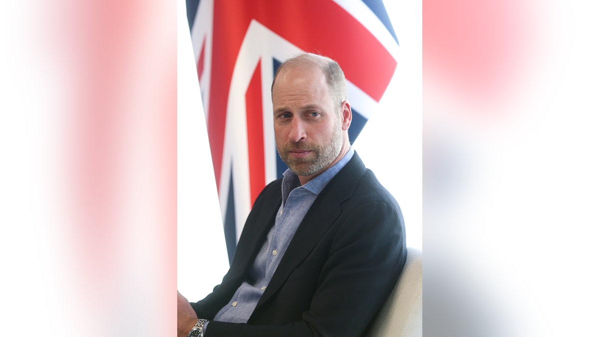 Prince William sitting in a suit in front of a Union Jack flag.