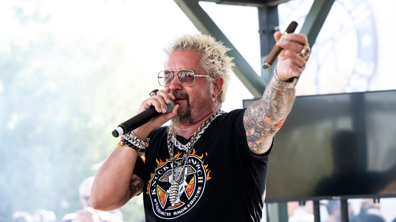 Guy Fieri holds a cigar in one hand and a microphone in another while speaking at an event