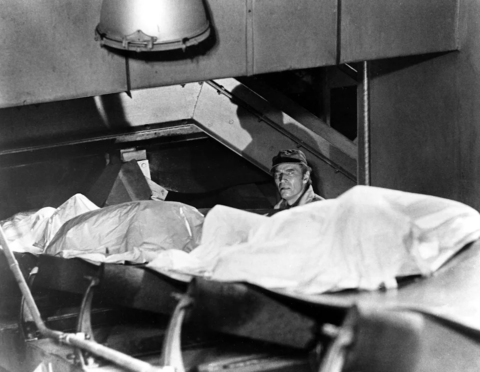 A person in military attire stands beside covered bodies on a ship during a historical scene, conveying a somber moment