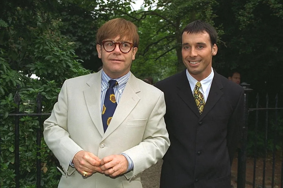 Two people in suits outdoors, walking side by side, smiling. One person wears glasses and a patterned tie; the other wears a plain tie