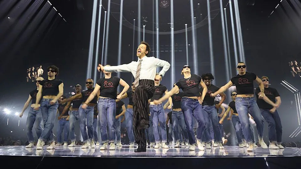 Harry Styles dances on stage at the Brit Awards. Wearing a shirt, tie and pinstripe trouser combination, he leads a group of dancers dressed in jeans and black t-shirts.