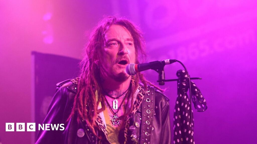 Ginger Wildheart performs with The Wildhearts at The 1865 in Southampton on 16 March 2025. He has long hair in dreadlocks, a short beard and moustache  and is wearing a leather jacket and an anarchy symbol and razor blade around his neck. He has musical notes tattooed along the base of his neck and there is a blue scarf with white polka dots tied to his microphone stand. He is singing into a microphone.