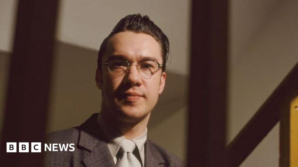 Mark Lamarr in glasses, a quiff, suit and tie in an official portrait photo taken in 1995