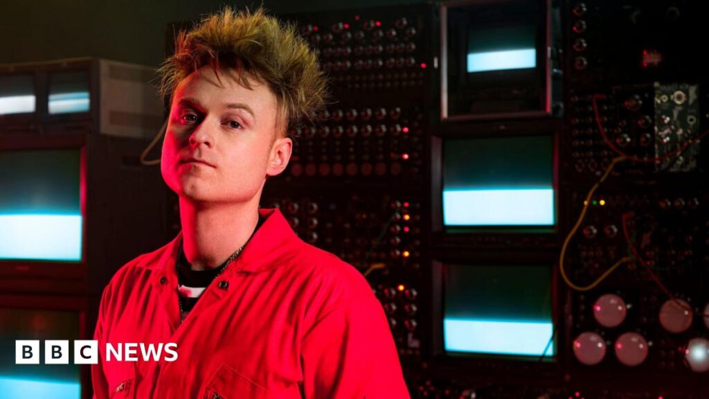 Look Mum No Computer, aka musician Sam Battle, stands in front of a bank of vintage synthesisers and TV monitors.