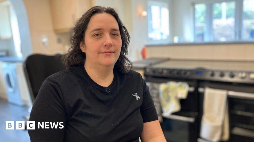 Susannah Thompson has long dark hair and a black t-shirt with a ribbon logo. She is sat in a kitchen.