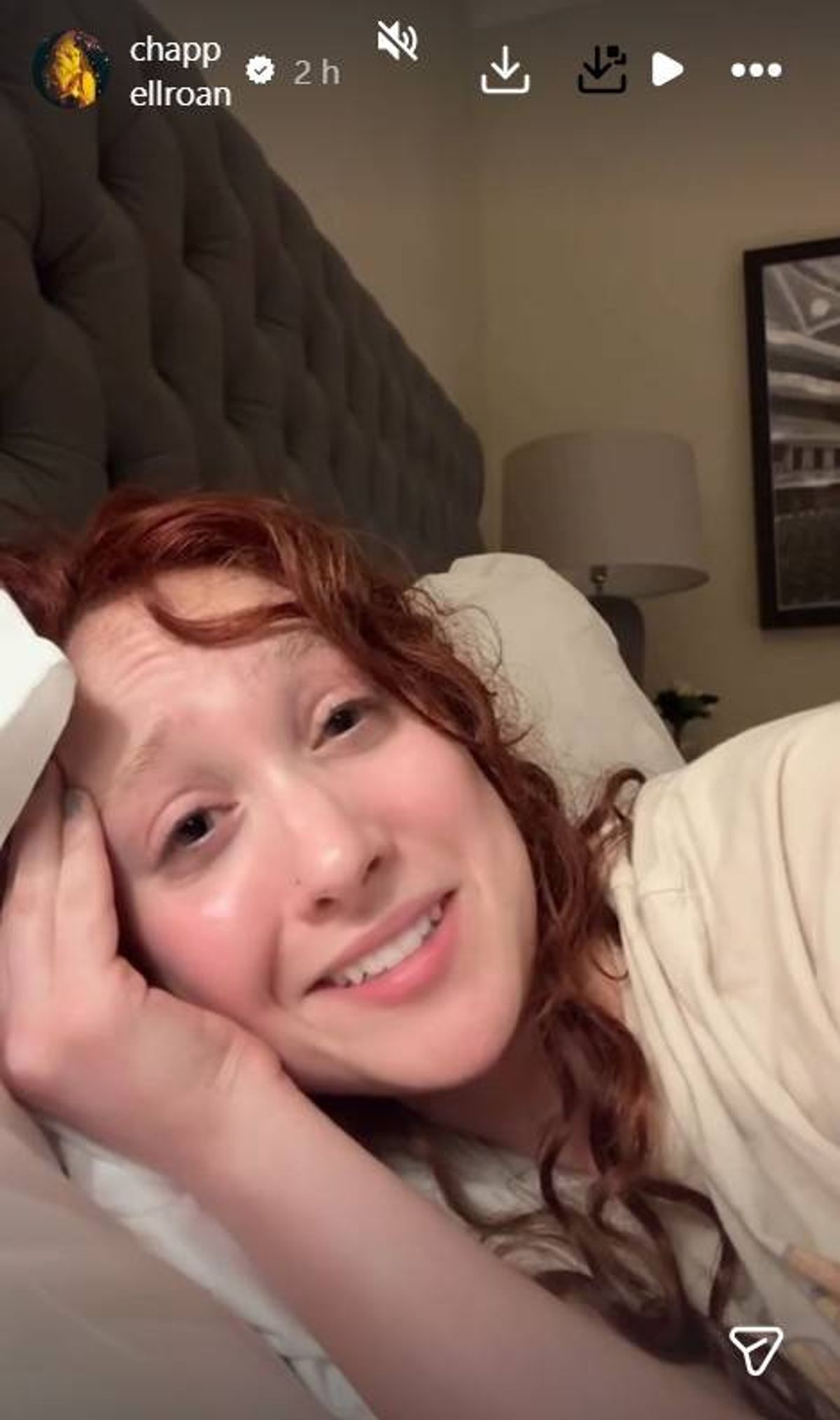 An individual with curly hair is reclining on a bed, their head resting on a hand, and they are smiling warmly while looking directly at the camera. The setting appears to be an indoor room, with a portion of a white pillow visible in the background.