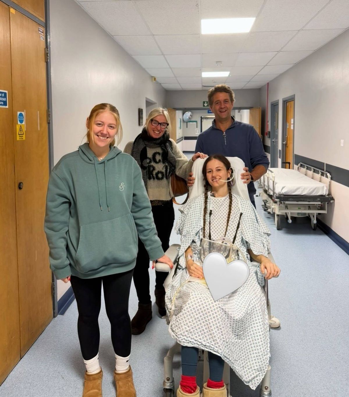 In a hospital corridor, a group of individuals is standing together. Among them, a person is seated in a hospital bed, accompanied by another individual standing beside her. Behind them, a hospital bed is positioned near the wall.