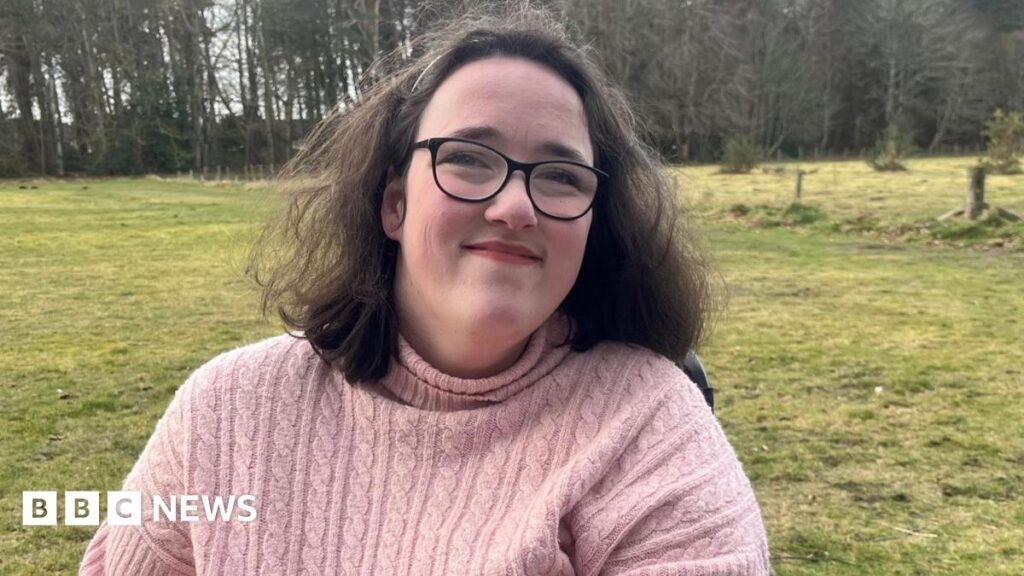 Iona Grant has dark hair down to her shoulders and is wearing a hair band and glasses. She is also wearing a pink woolly jumper. She is in a wheelchair in the outdoors. There is grass and woodland behind her.