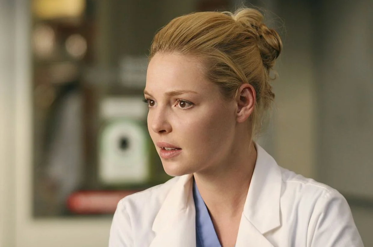 A woman with blonde hair in a neat bun, wearing a white lab coat and blue scrubs, looks to the side with a thoughtful expression in a medical setting.