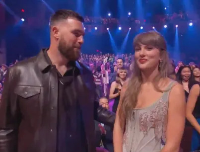 A celebrity couple stands together at an event; the man wears a leather jacket and the woman is in an embellished dress, surrounded by a cheering crowd