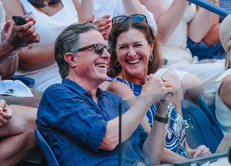 A couple smiles and holds hands while seated in a crowd at an outdoor event, surrounded by other attendees