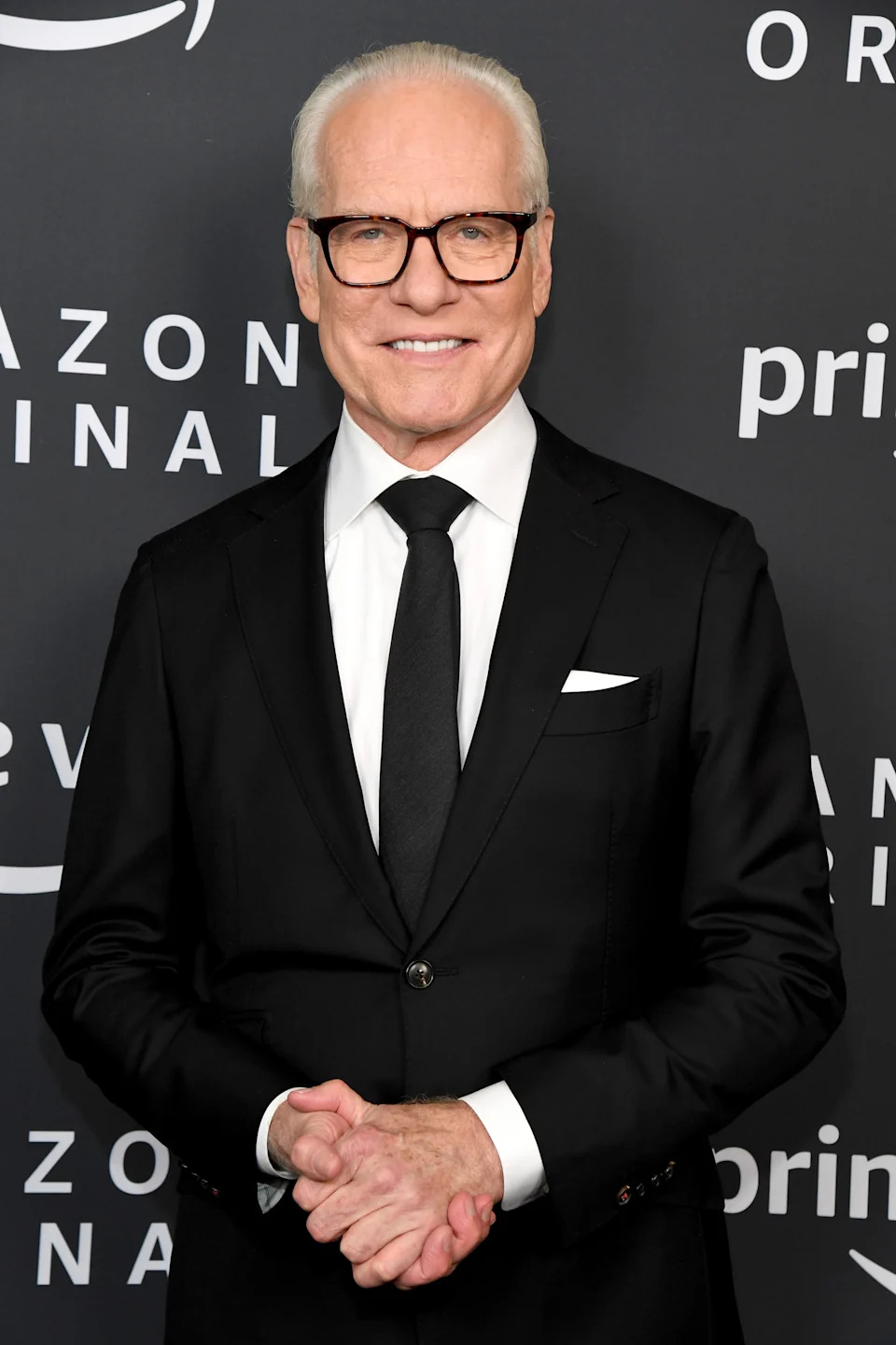 Tim Gunn in tailored suit and tie on the red carpet, smiling in front of a backdrop with logos