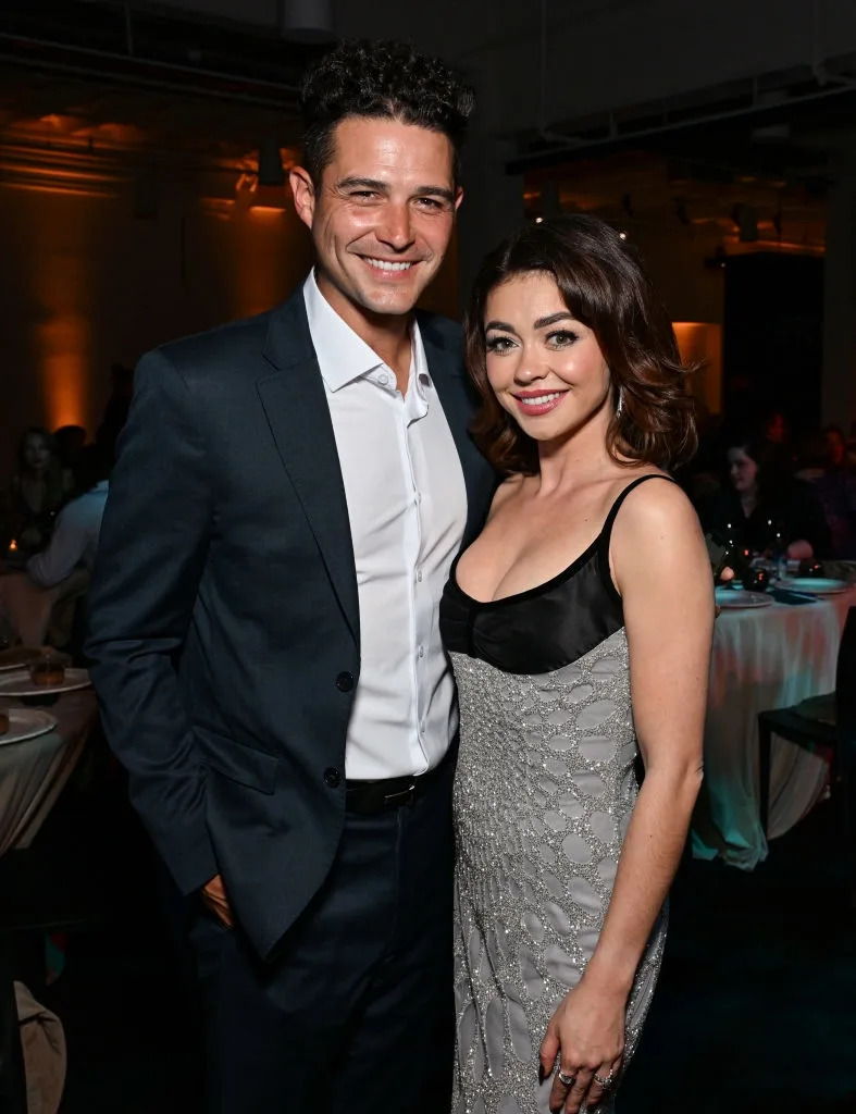 Two people are smiling, standing together at an event. One person wears a suit, and the other wears an elegant dress with a shimmering texture