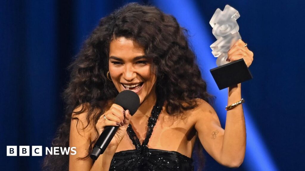 Olivia Dean wears a black sparkly strappy dress. She is dark brown hair and wears hoop earings. She is standing on stage accepting a silver trophy.