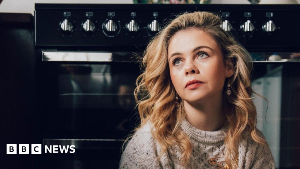 Vogue: Hairdresser says working with Derry Girls actor Saoirse-Monica Jackson ‘surreal’ A woman stands in a white shirt and black tie with a red and navy tartan blazer. She has long brown hair and is wearing red lipstick.