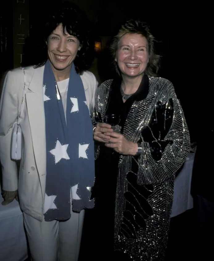 Two people smiling at an event; one in a star-patterned scarf and white suit, the other in a sequined jacket, holding a drink