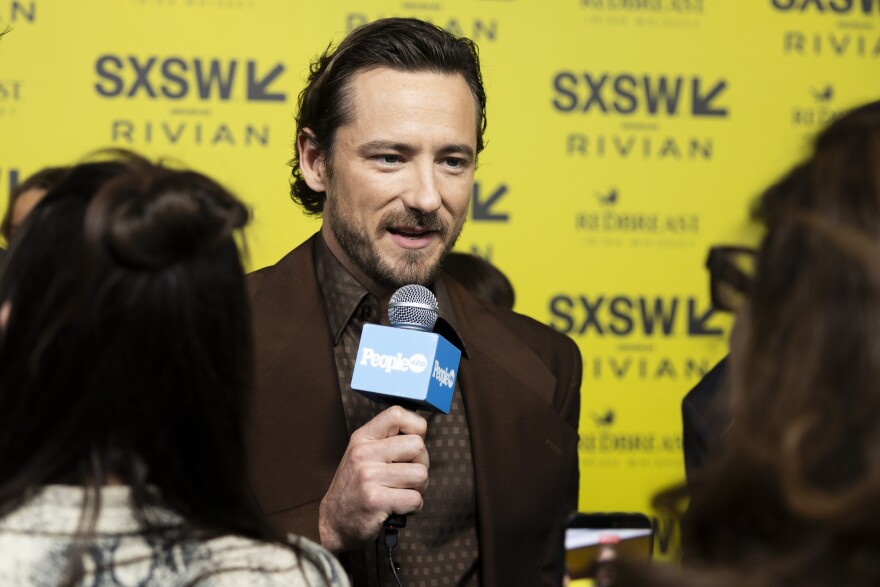 Lewis Pullman speaks into a microphone in front of a yellow backdrop covered in SXSW logos.