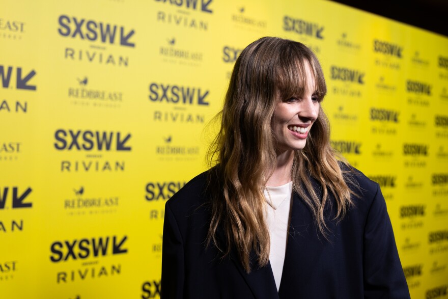 Maya Hawke stands, smiling, in front of a yellow backdrop covered in SXSW logos.