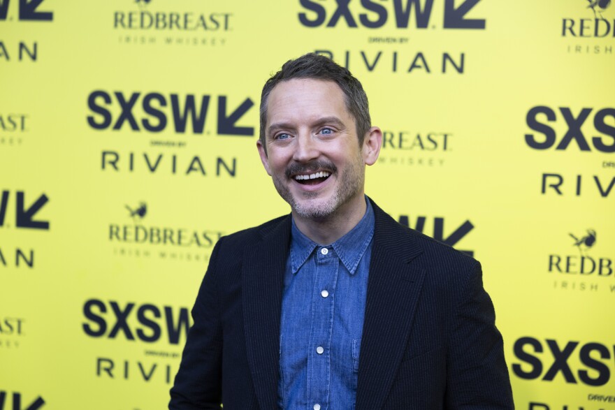 Elijah Wood smiles in front of a yellow backdrop with SXSW logos on it.