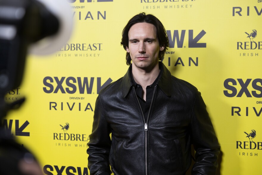 Cory Michael Smith poses for photos in front of a yellow backdrop with SXSW logos on it. 
