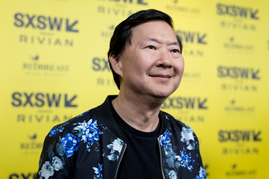 Ken Jeong smiles in front of a yellow backdrop with SXSW logos on it.