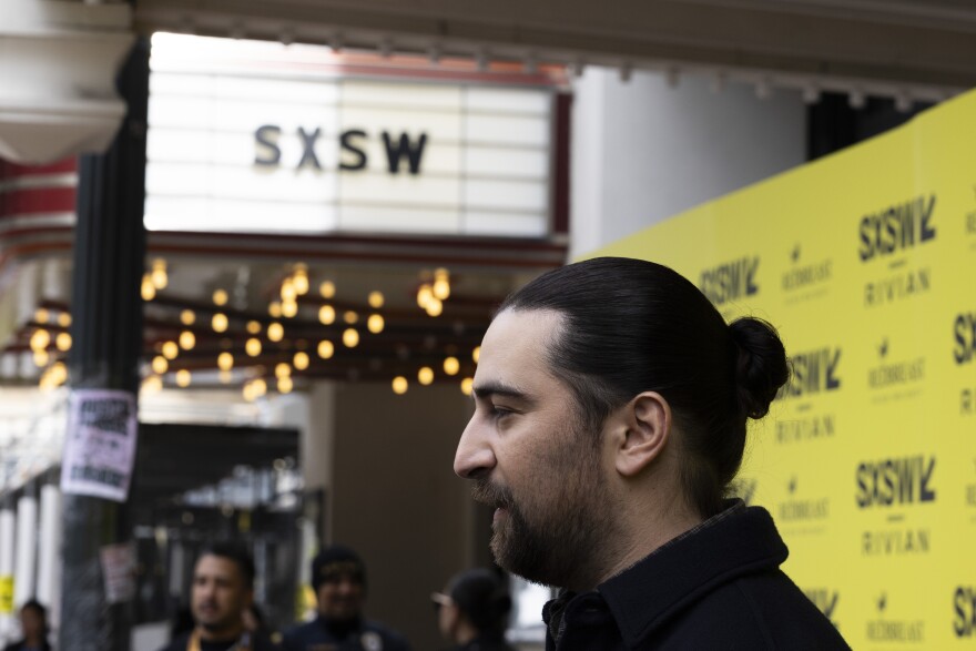 Noah Kahan looks to the left at cameras while standing in front of a yellow backdrop with SXSW logos.