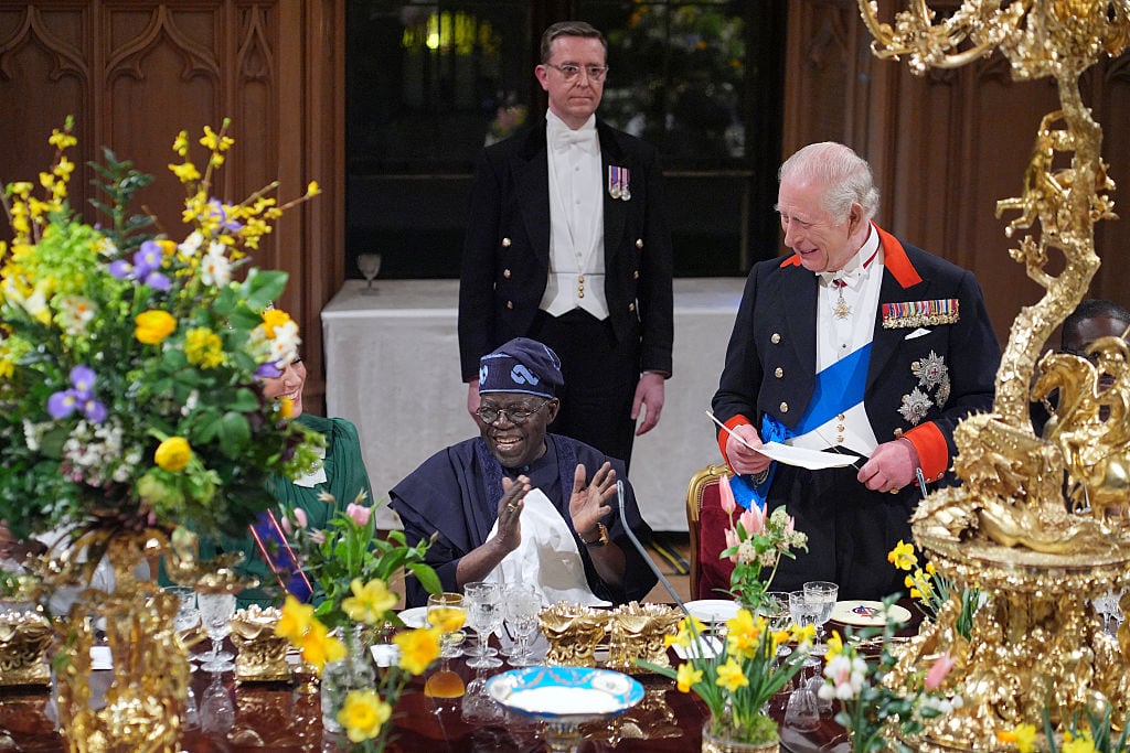 A person standing and holding papers looks toward a person seated at an elaborately set dining table, which displays large bouquets of spring flowers and gold-plated decor.