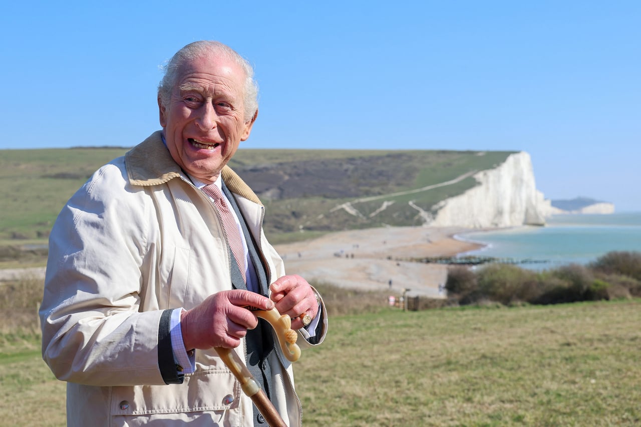 A person holding a walking stick smiles as they stand along a coastal path with a chalk cliff face and the shore in the background.